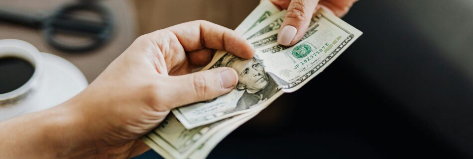 A close-up image of hands exchanging US dollar bills, symbolizing financial transaction or payment.